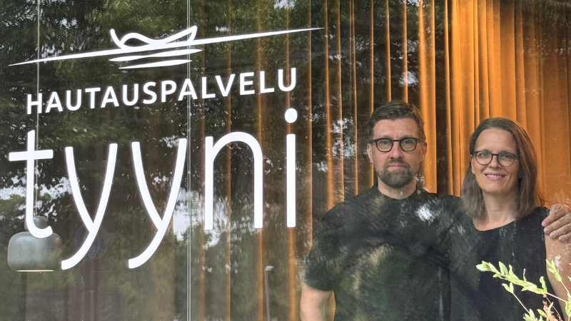 Hautauspalvelu Tyynin yrittäjät Susanna ja Jouni Lahtinen seisovat liikkeensä ikkunan edessä, jossa näkyy yrityksen logo